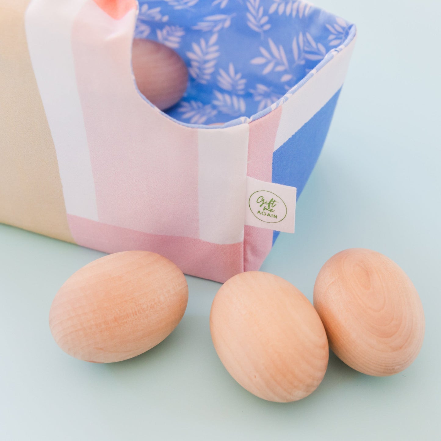 Reusable fabric Easter basket with pastel brushstroke exterior and blue leaf print lining, filled with wooden eggs. In front of the basket are also 3 wooden eggs.