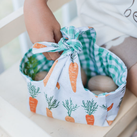 A child holds a small, reusable Easter basket made from fabric with a cute carrot print on the outside and green gingham lining. The basket is tied at the top and contains a few wooden eggs.
