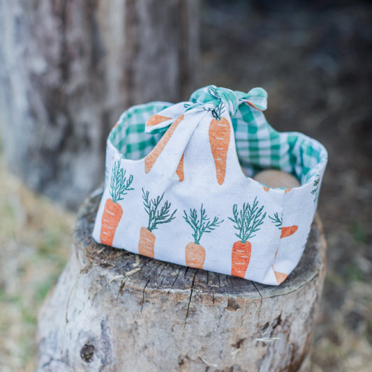 A reusable, reversible Easter basket made from fabric with a carrot print exterior and green gingham lining, displayed outdoors on a tree stump.