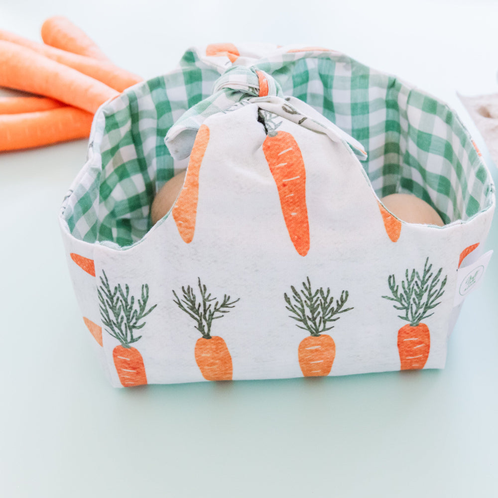 A reusable Easter basket made from fabric with a cute carrot print on the outside and green gingham lining. The basket is tied at the top and contains a few wooden eggs. Next to the basket are some carrots.