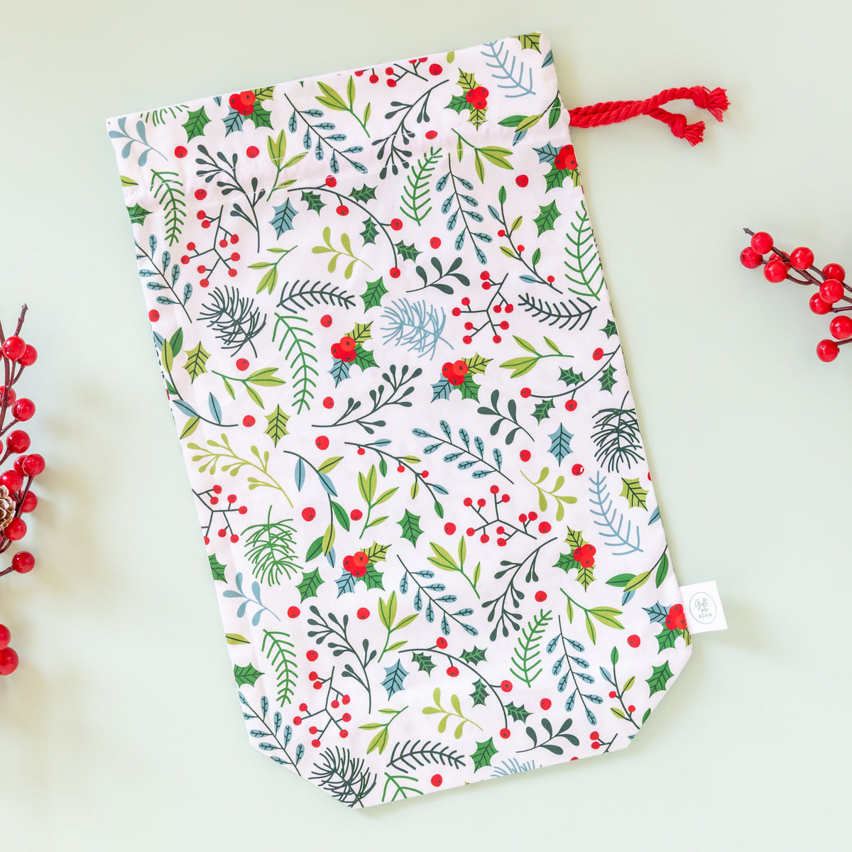 A fabric drawstring pouch with a festive Christmas botanical print, featuring green and red foliage on a white background, styled with red berry branches on a pale mint green surface.