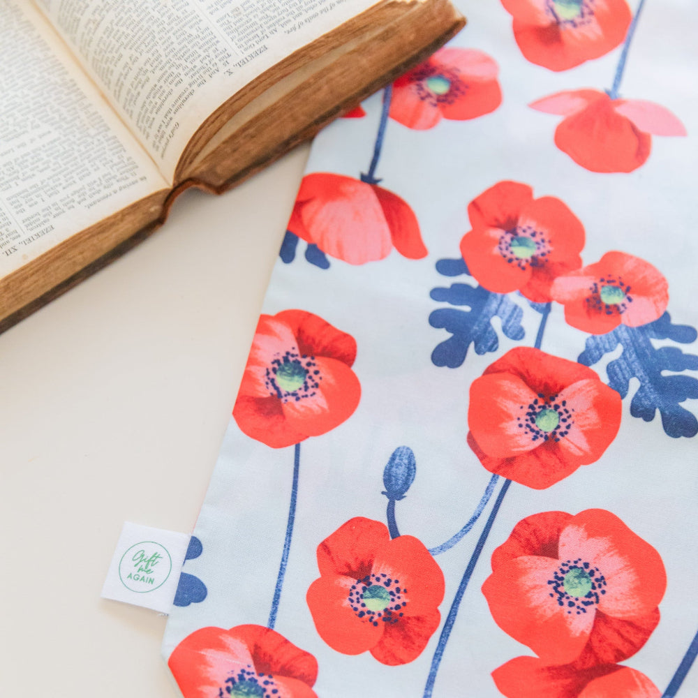 A fabric gift bag laid flat with red poppies against a light blue background with a Gift Me Again tag