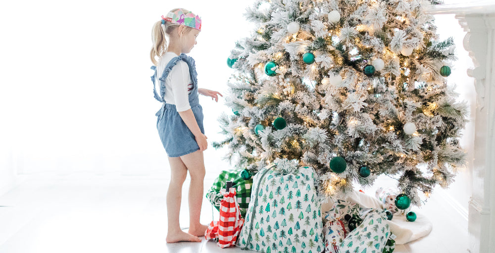 Child standing next to a decorated Christmas tree with presents wrapped in fabric gift bags underneath.