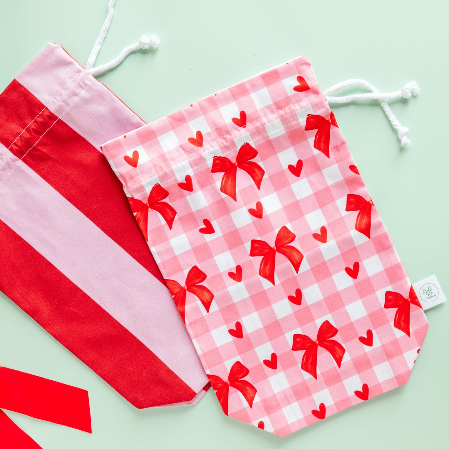 Two drawstring gift bags, one red and white striped, the other with a pink checkered pattern and red bows, on a light green background.