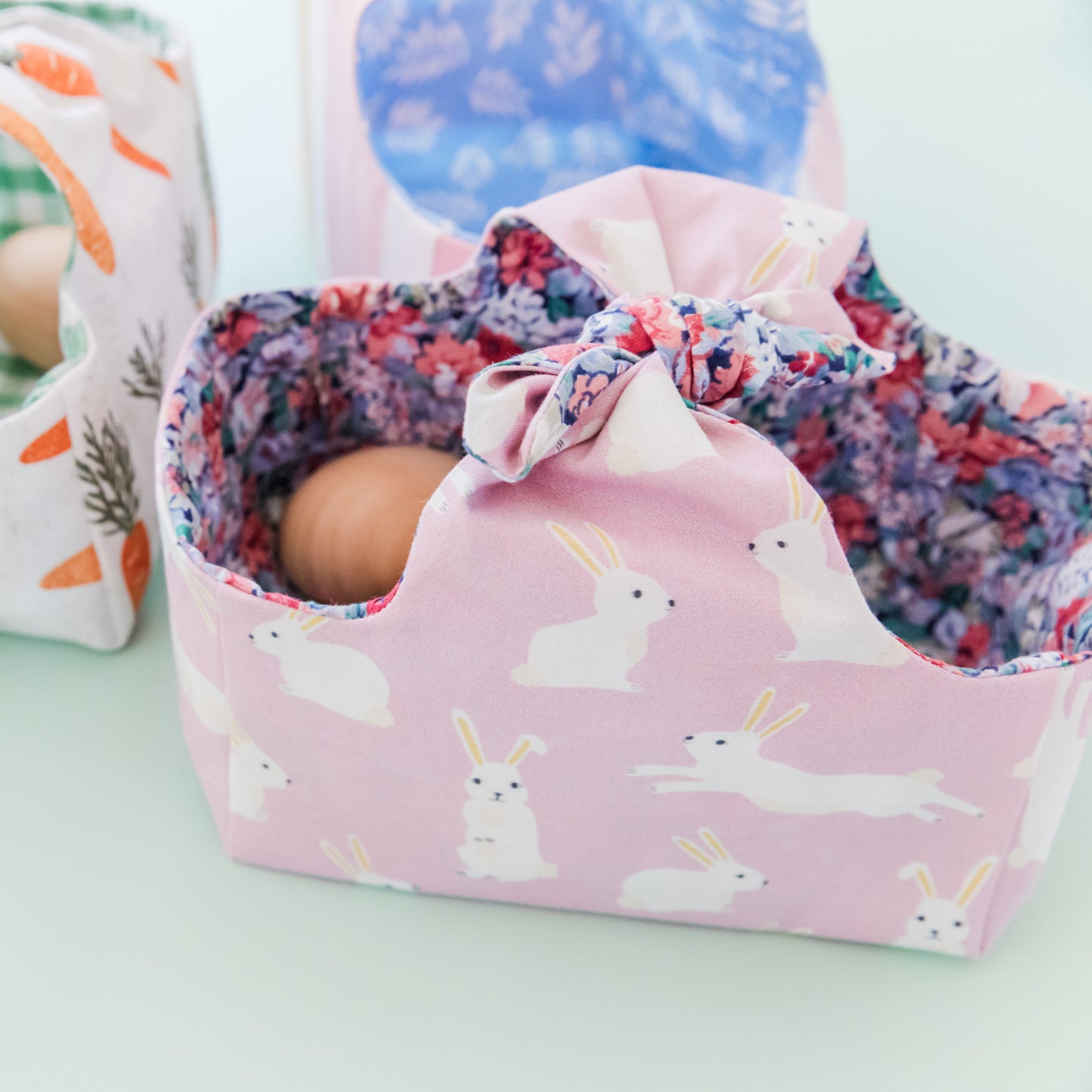 A fabric Easter basket in a Lilac fabric featuring white rabbits and lined with a floral fabric. Inside the basket are some wooden eggs.