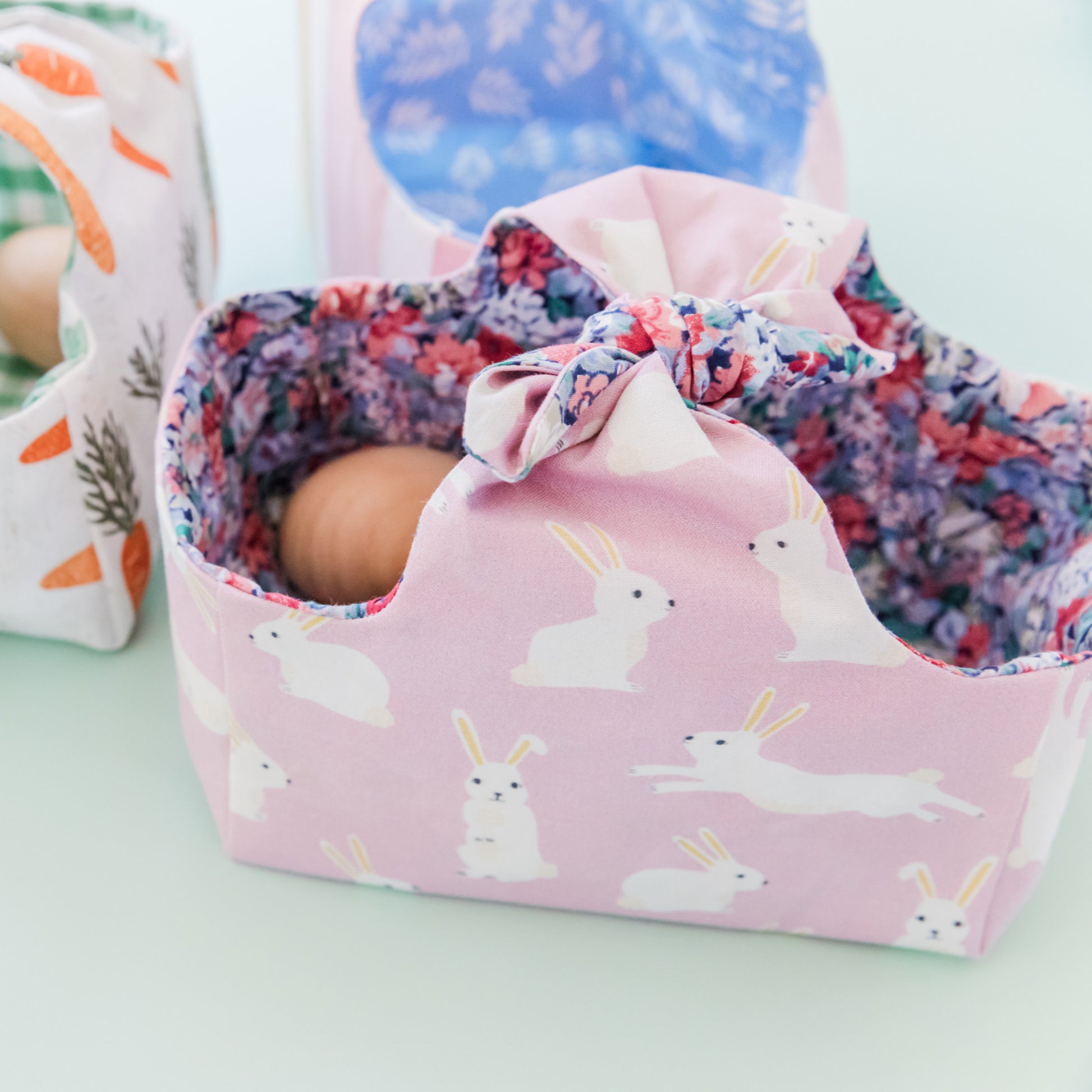 A fabric Easter basket in a Lilac fabric featuring white rabbits and lined with a floral fabric. Inside the basket are some wooden eggs.