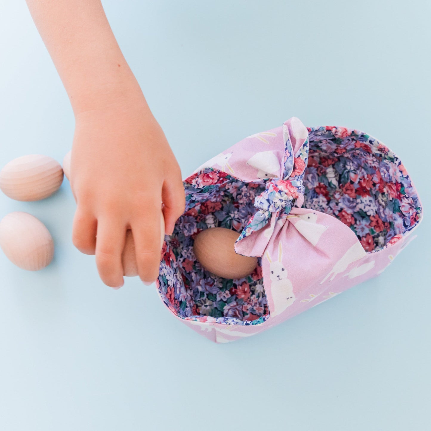 A fabric Easter basket in a Lilac fabric featuring white rabbits and lined with a floral fabric. A child's hand is placing wooden eggs inside the basket.