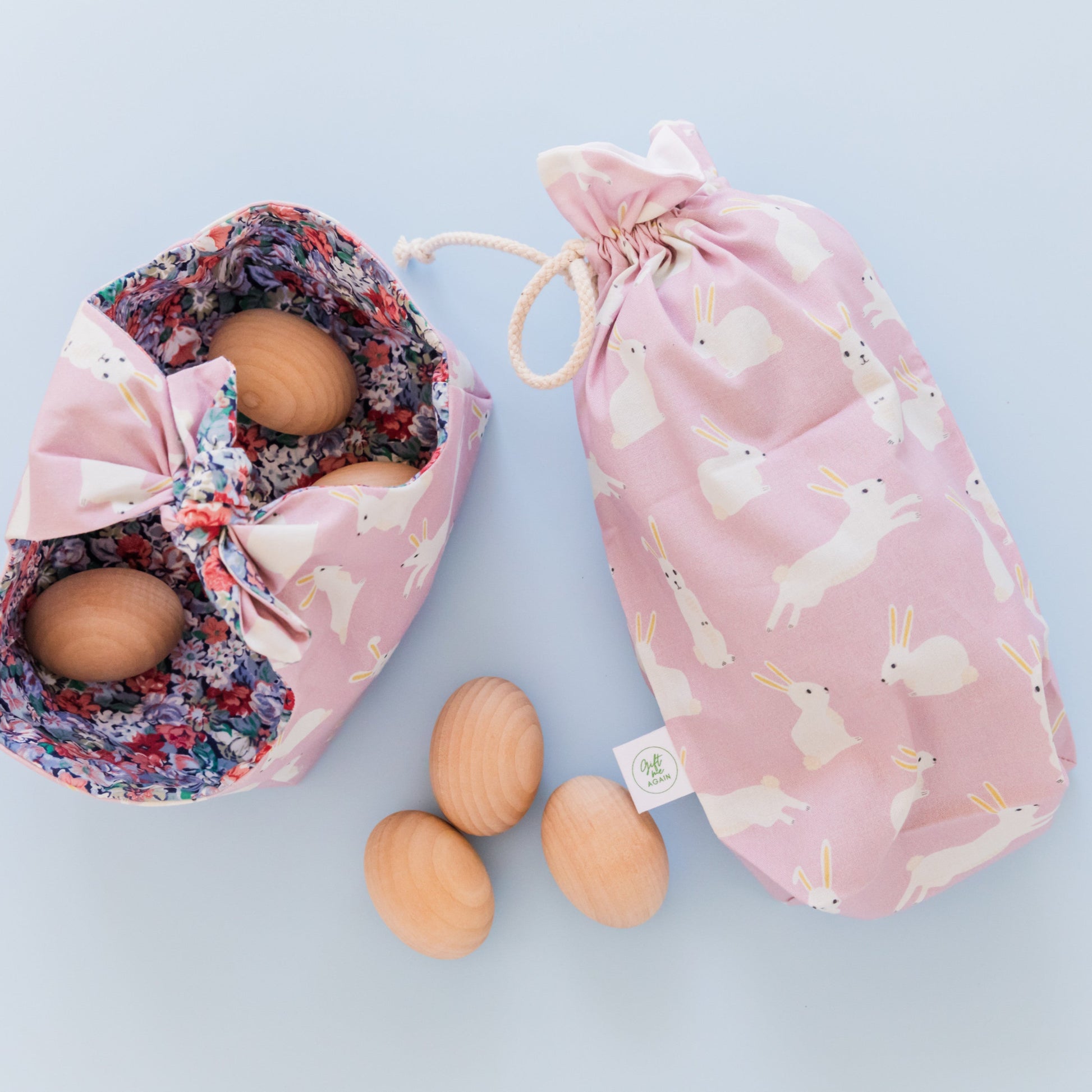 A fabric Easter basket in a Lilac fabric featuring white rabbits and lined with a floral fabric. The basket sits next to a fabric gift bag in the same lilac bunnies print. Inside the basket are some wooden eggs.