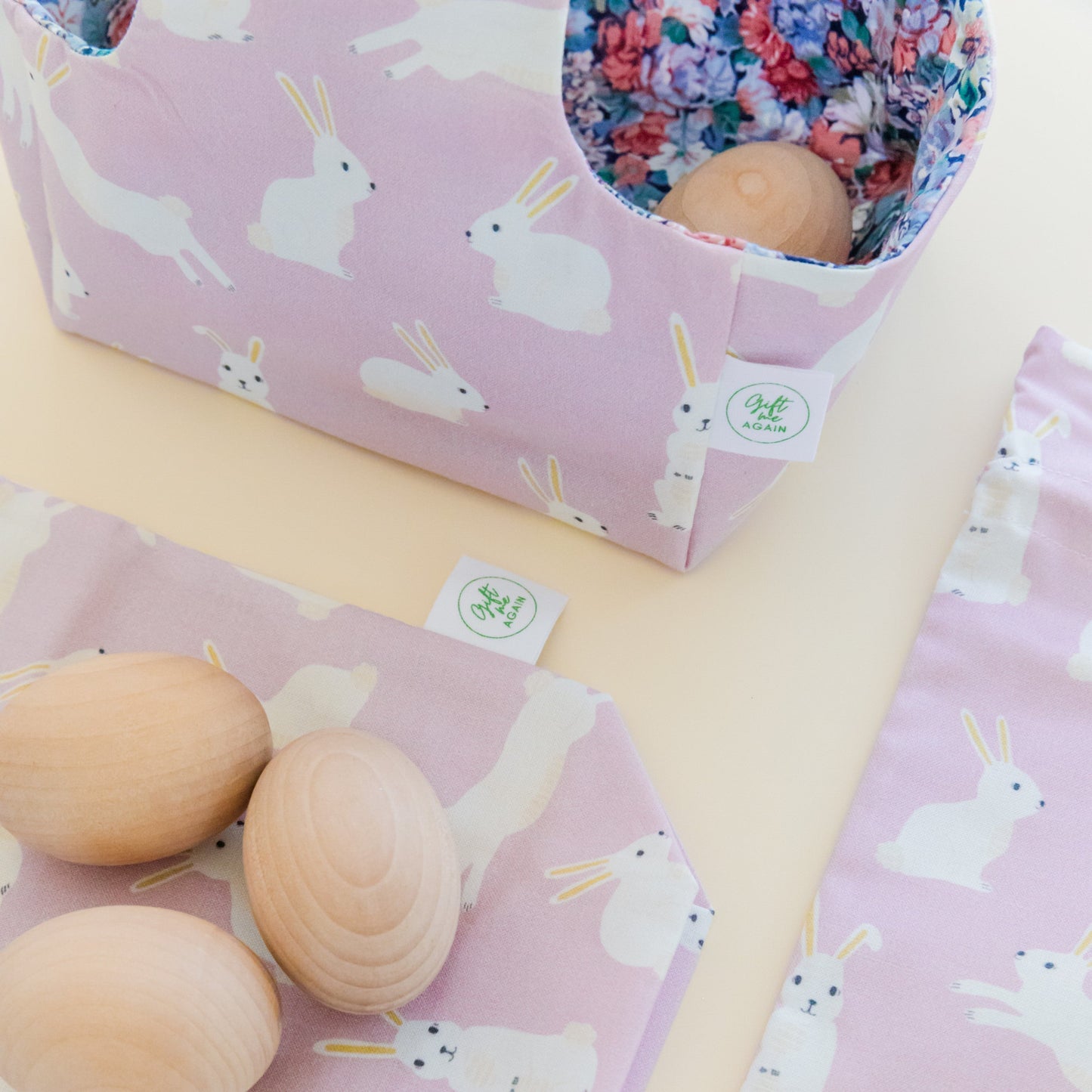 Two fabric gift bags in a lilac bunnies print sit flat. On top of the gift bags are some wooden eggs and behind is a fabric Easter Basket in the same print.