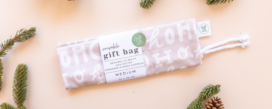 Neutral colour reusable gift bag in print with Ho Ho text, against a light background with pine cones.