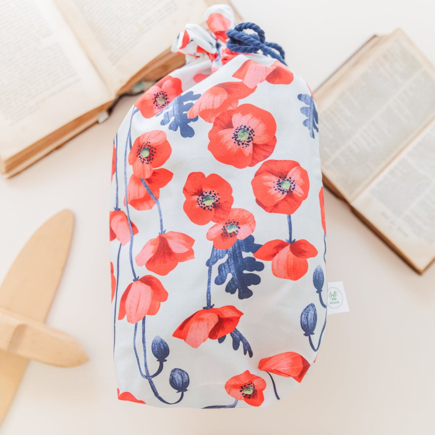 Reusable fabric gift bag featuring a vibrant red poppy floral print, handmade by Gift Me Again. Displayed beside open books for a soft, natural look.