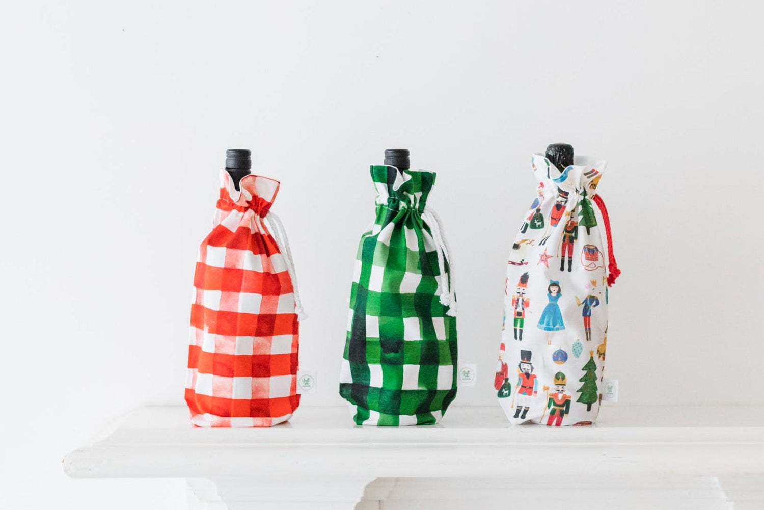 Three reusable Christmas bottle gift bags displayed on a white mantel, featuring red gingham, green gingham, and a festive Nutcracker print. Eco-friendly fabric bags handmade by Gift Me Again, perfect for wrapping wine or olive oil sustainably.
