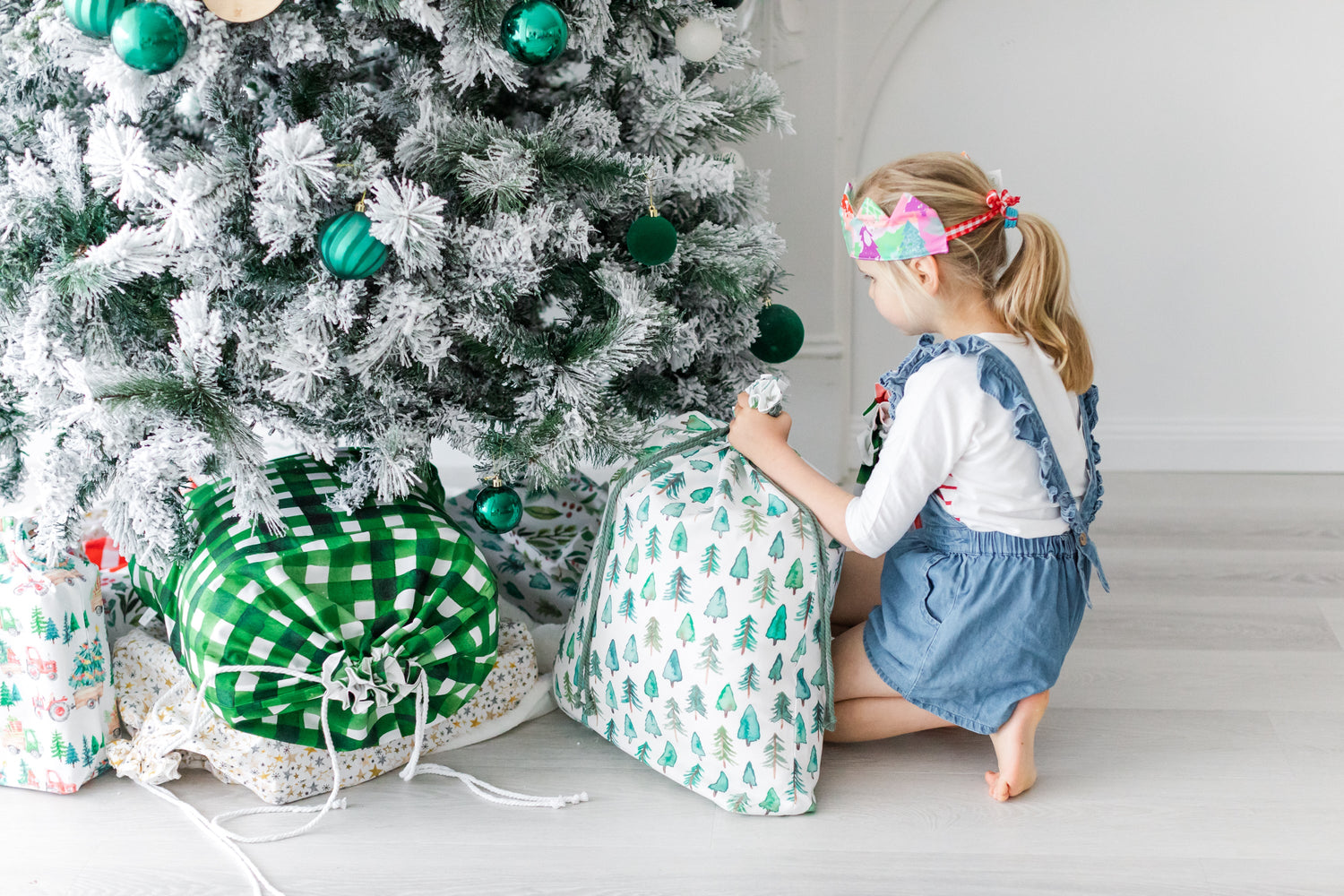 A girl wearing a crown places an extra large gift sack under a Christmas tree. 