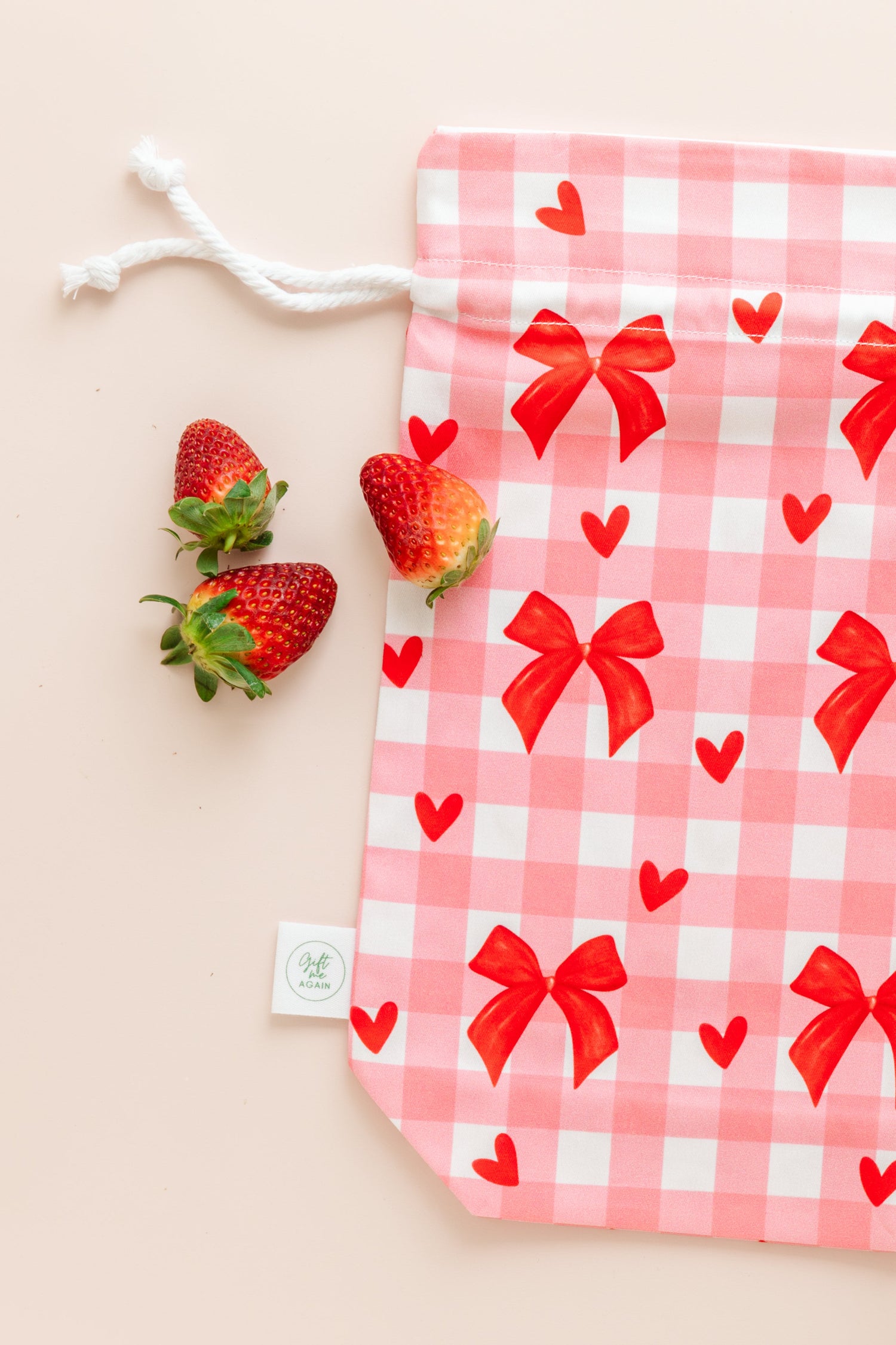 Pink and white checked gift bag with bows and hearts surrounded by strawberries on a beige background