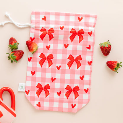 Pink checkered drawstring gift bag with red bows and hearts, surrounded by strawberries on a beige background