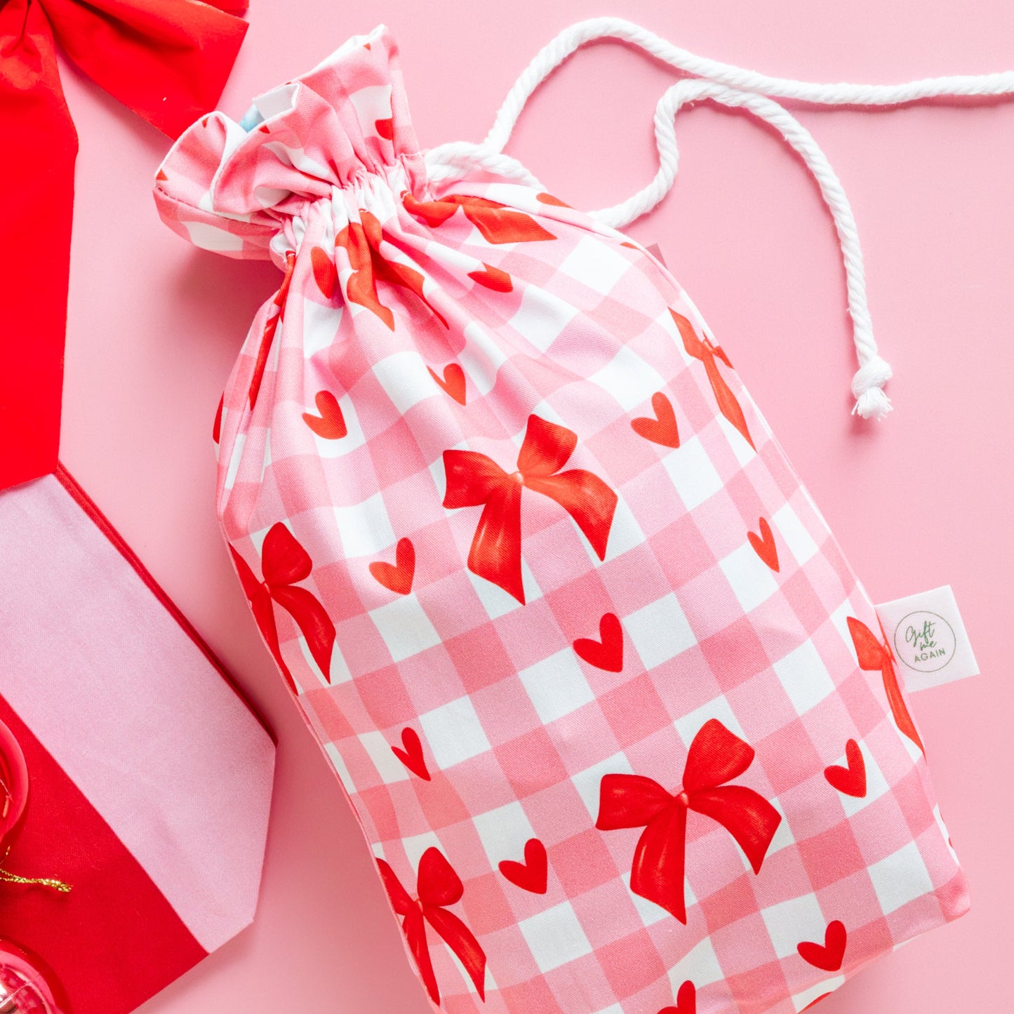 Drawstring gift bag with red bows and hearts on a pink checkered pattern, placed on a pink background.